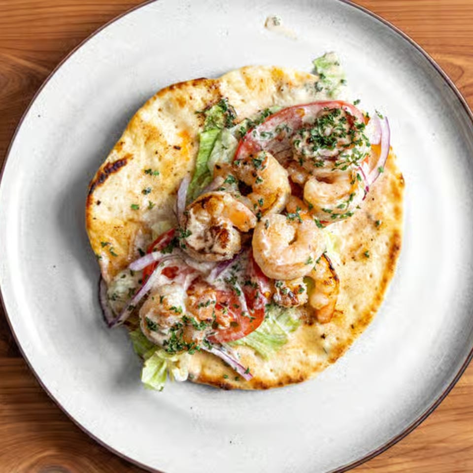 Grilled shrimp flatbread with lettuce, tomatoes, red onions, and a creamy sauce on a white plate.