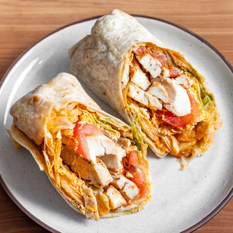 Two halves of a chicken wrap, filled with chicken, lettuce, tomatoes, and cheese, on a white plate.